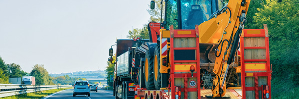 A heavy truck on a tree-lined highway pulls a trailer of large construction equipment     